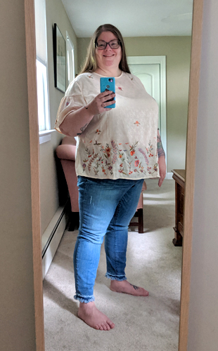 image of me standing in a full-length mirror with my hair down, wearing grey-framed glasses, a pale pink top embroidered with flowers, and distress medium-wash jeans