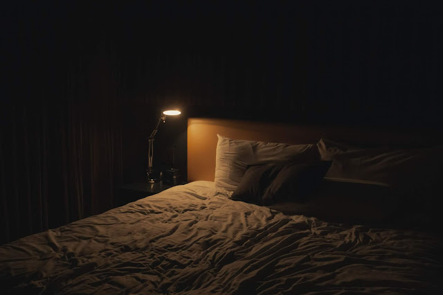A bedroom at night. A bed partially illuminated by a brassy gold lamp.