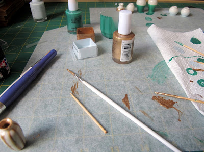 White dolls' house vase painted with gold stripes, on a workbench with bottles of nail varnish and other, dipped, vases in the background.