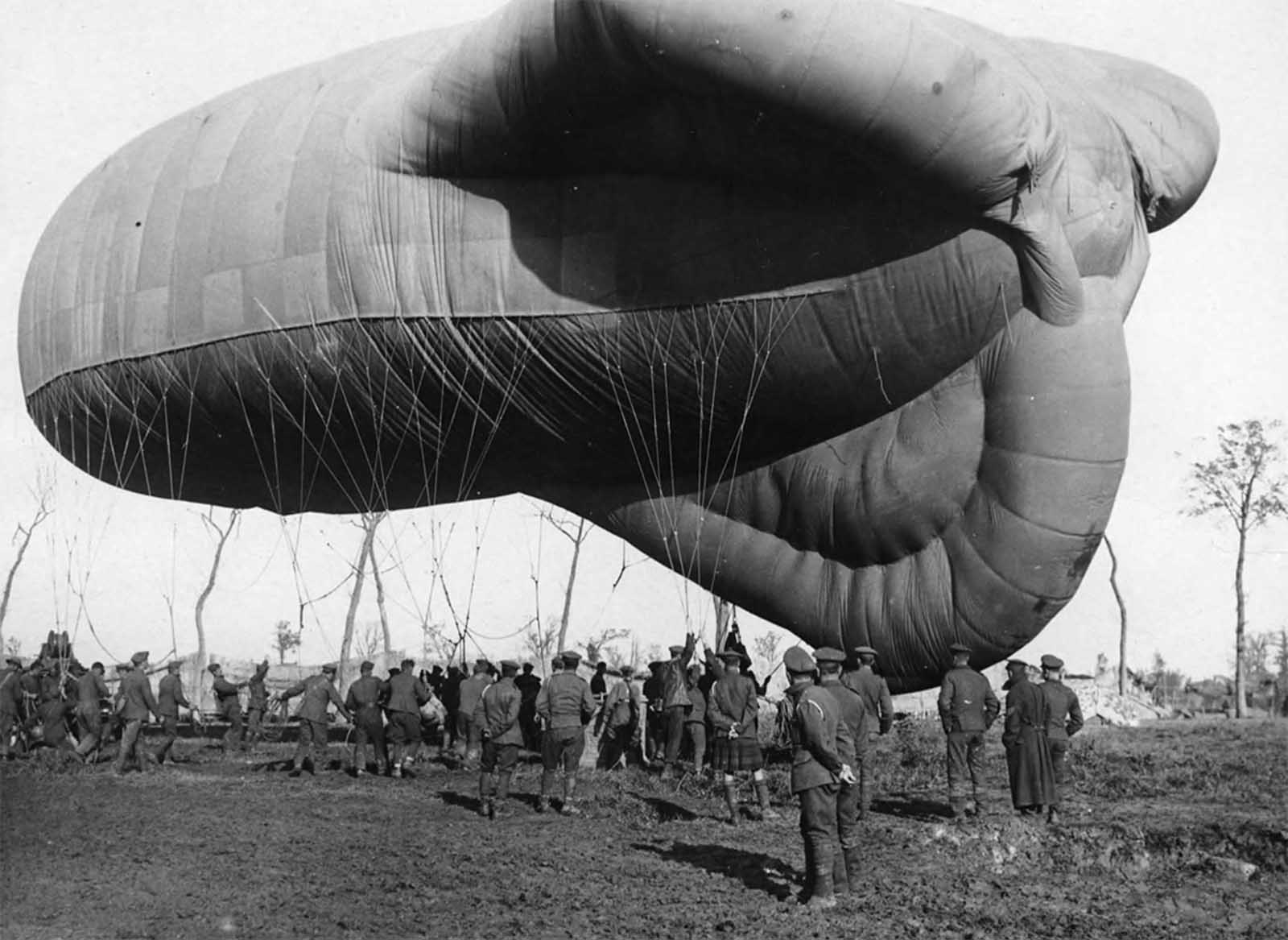 A returning observation balloon. A small army of men, dwarfed by the balloon, are controlling its descent with a multitude of ropes. The basket attached to the balloon, with space for two people, can be seen sitting on the ground. Frequently a target for gunfire, those conducting observations in these balloons were required to wear parachutes for a swift descent if necessary.