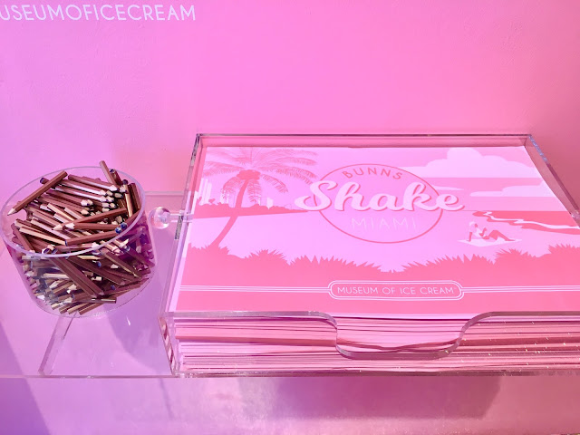 A clear plastic cup filled with mall golden pencils next to a receptacle filled with pink paper placemats