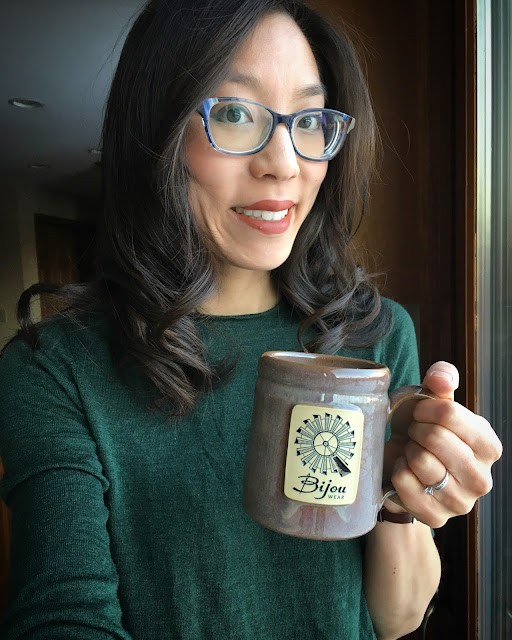 [Image of a smiling tan skin bespectacled Asian woman wearing a forest green sweater and holding a tan glazed ceramic mug. The mug is filled with hot milk, a splash of coffee, and a drizzle of honey. It features a rectangular medallion with a windmill and text Bijou Wear. Woman’s dark brown hair is curled because she wanted to curl it today.]