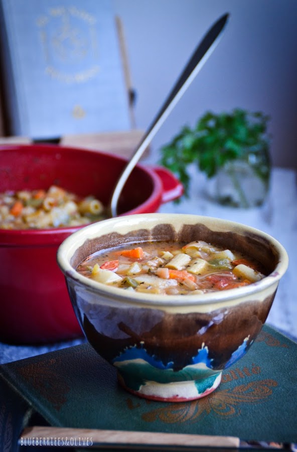 Olla cerámica roja llena con hoja laurel, bol cerámica con sopa, paño lino gris, libro cocina del mundo, cuchara mango madera,hojas perejil fresco
