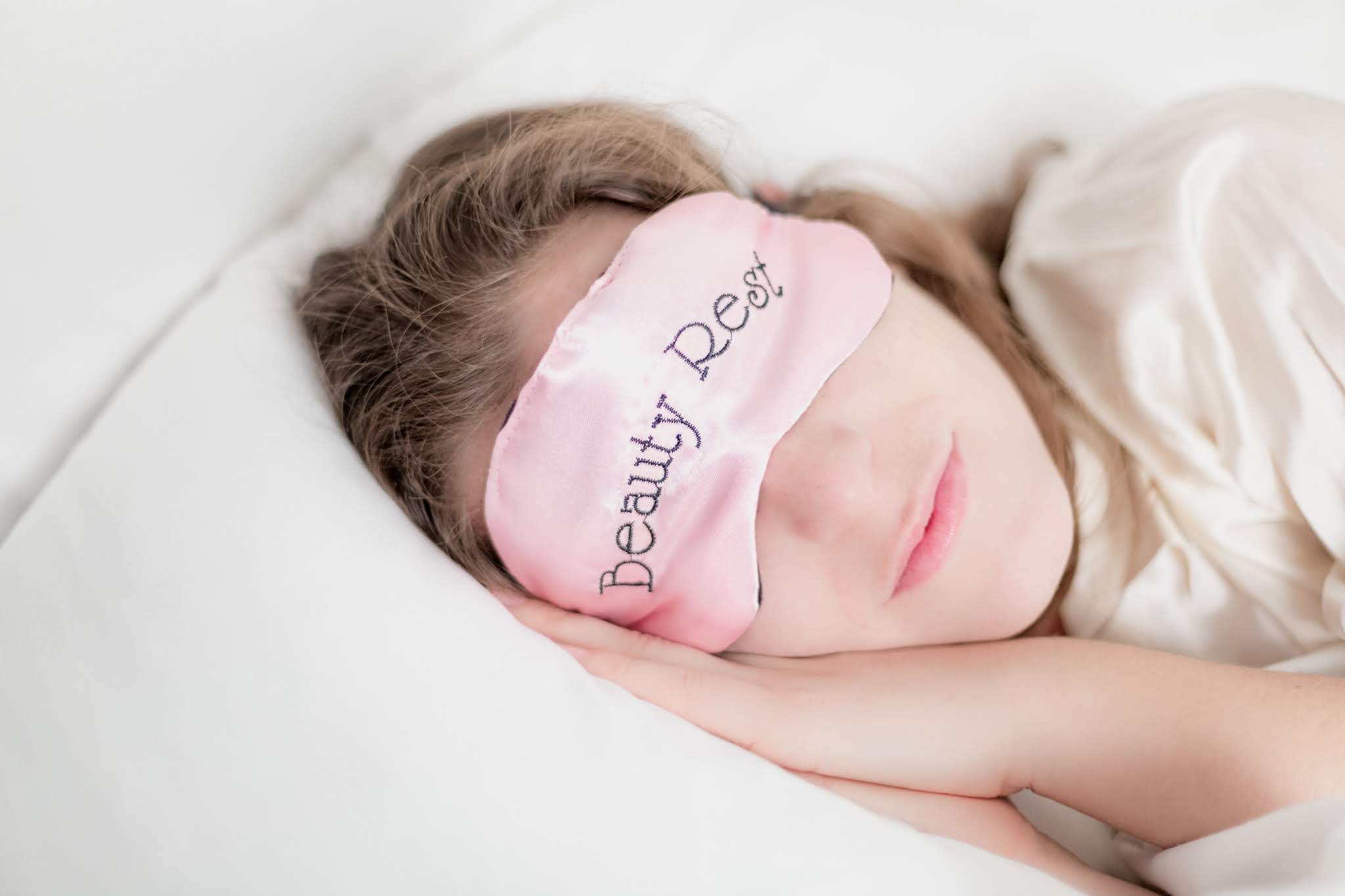 A woman rests her head on a pillow and wears a pink satin mask that states: "Beauty Rest", allowing her to catch a little extra sleep.