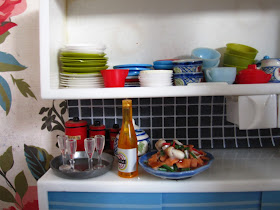 One-twelfth scale modern miniature kitchen dresser filled with mismatched crockery and holding a tray of champagne glasses, a bottle of sparking wine and a platter full of seafood.