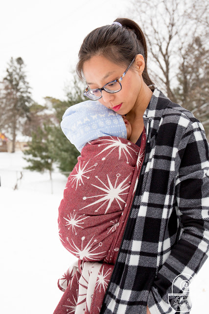 Image of a tan skin bespectacled Asian woman with dark brown hair in a high pony tail wearing a sleeping toddler in a maroon and ecru retro star patterned woven wrap carrier on her front underneath a black and white plaid fleece jacket. They're outside in a winter wonderland with evergreens in the background.