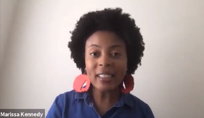 Young African American woman seated for the camera. She is wearing a blue shirt and large red earrings.
