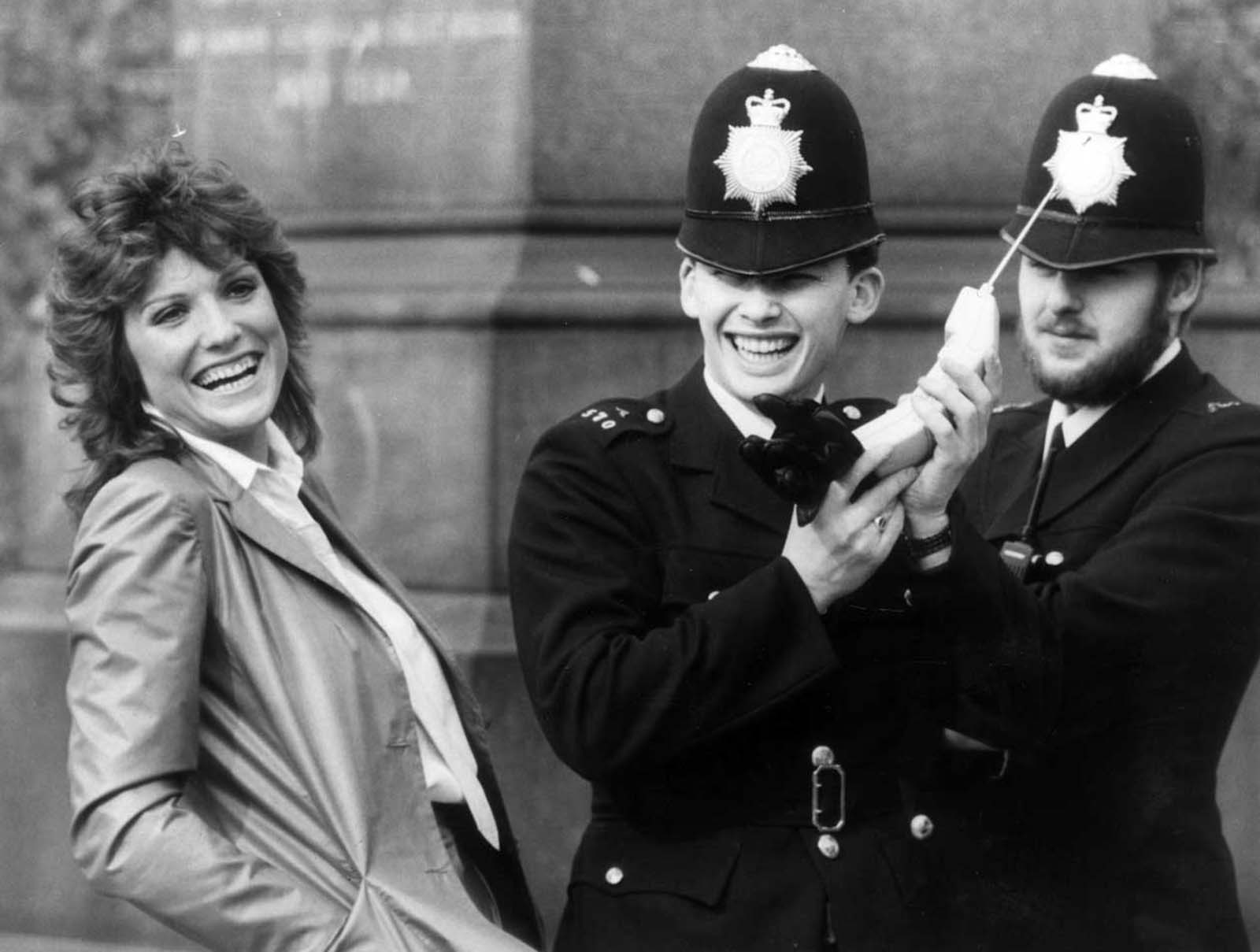British actress Suzanne Danielle sharing a joke with two policemen while they examine one of the then-newly legal mobile phones in the United Kingdom. 1983.