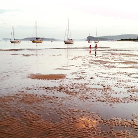 Beach at sunset with the tide out. Two people are paddling in the distance, and behind them are a number of moored yachts.