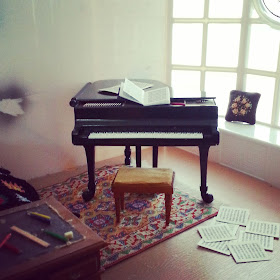 One-twelfth scale modern miniature piano studio with a grand piano in front of a bay window. On the floor are strewn several pages of music.
