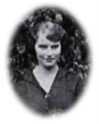 A smiling young white woman with 1920s style clothes, hair, and makeup standing in front of some shrubbery