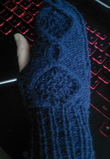 The back of the hand of a cabled mitten.  The thumb stitches are live and on a dpn. Background is a computer keyboard.