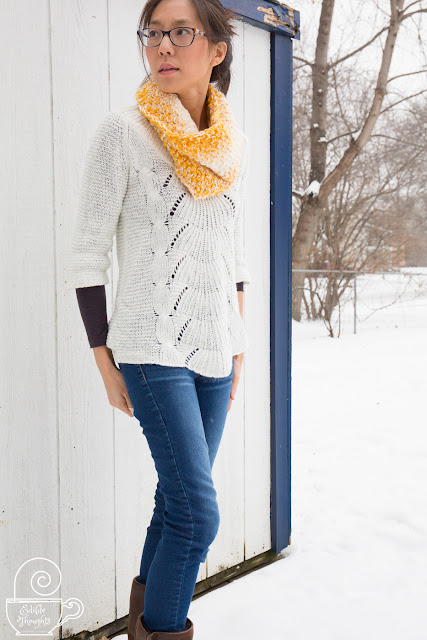 Image of a tan skin bespectacled Asian woman looking to her right. She's wearing a yellow and white crocheted cowl with a white sweater over a heathered charcol thermal long-sleeve over skinny jeans tucked into brown boots. She's in front of a worn white shed in the snow with bare branched trees in the background.