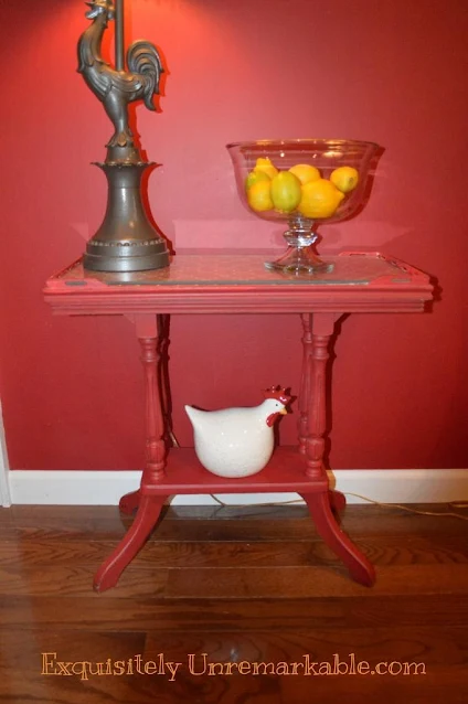 A red painted table with rooster lamp, lemons in a bowl and small red ceramic chicken on bottom shelf