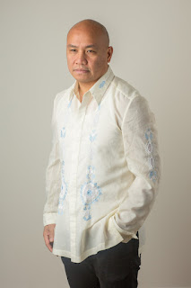 Photo of Patrick Rosal. Image of a bald Asian man with a neutral expression standing in front of a beige wall. He wears a cream colored long sleeve button up shirt with light blue embroidery down the front.