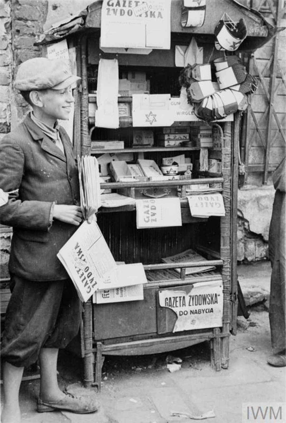 A young and cheerful seller of newspapers and armbands running his stall in the street of the ghetto (possibly Muranowski Square). The title of the newspaper for sale is 