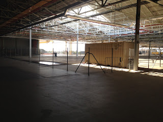 A view of an empty industrial building with concrete floors. The roof framework is exposed. A section of roof is open to the weather. A yellow shipping container stands in the centre of a fenced-off square. The entrance is in the distance. The light can be seen in contrast to the shadows of the inside of the building. A shallow pool of water has gathered underneath the open section of roof after recent rains.