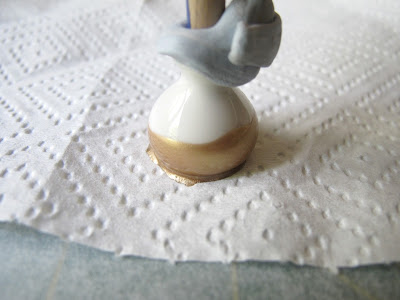White dolls' house miniature vase, with the end of a paint brush blue tacked into it, half-dipped with gold nail varnish, on a paper towel.