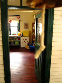 View through the front door of a miniature dolls' house school, showing the kitchen with a selection of drinks arranged.