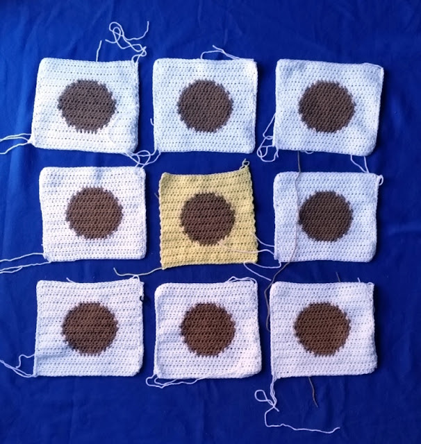 Nine squares are arranged on a royal blue background in a 3 by 3 arrangement. All of the squares are white with tan circles in the middle except for the centre square which is yellow with a tan circle.  The squares still have loose ends of yarn at the corners.