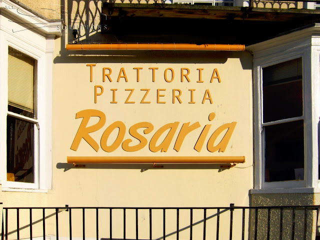 Photograph of light orange flat cut letters with 'Trattoria Pizzeria' in a sans font and 'Rosaria' in a brush style font. Above and below the letters are two mounted bars, in the same colour as the text with lights inside for a bright fascia at night.