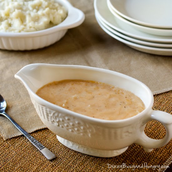 Side view of homemade gravy in a white gravy boat with a white bowl of mashed potatoes and a wooden spoon in the background. 