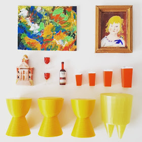 Flat lay of one-twelfth scale modern miniature items in colours of red, orange and yellow including paintings, Philippe Starck Prince Aha and Bubu stools, retro kitchen canisters and a bottle and glasses of red wine.