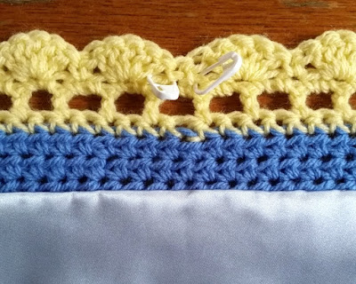The reverse edge of the baby blanket arranged in horizontal strips of colour. Brown across the top is the brown worktop. Yellow scalloped crocheted edging joined to the blue crocheted blanket by a row of yellow filet crochet.  The blue crochet is fabric made with half treble stitches (Australian/UK terms).  The pale blue satin edge is hem stitched to the back of the blanket along a row of blue half trebles.