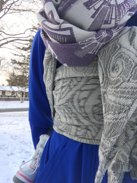 Image of a woman's torso featuring a gray botanical patterned woven wrap carrier spread across her chest over a blue coat. She's wearing a toddler on her back who has on white snow boots. They're outside on a winter day.]