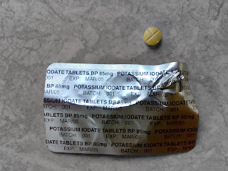 Front side of silver coloured blister pack containing six 85mg Potassium Iodate tablets in blister pack. One tablet has been removed from pack showing its cross-shaped grove along one side. The tablet has begun to yellow with age.