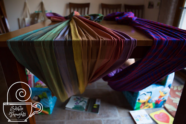 Image from the far end of two wrap hammocks with happy children inside and books scattered around the floor. In the background are chairs against the wall and bags hung on the wall. White watermark in bottom left corner of a steaming mug with a needle horizontal along the rim and curly text Edible Thoughts copyright symbol Stephanie Lu Sinclair.]