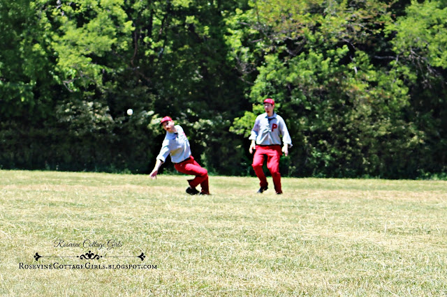 Two men in a grassy field playing outfield in a vintage baseball game.  They are wearing red slacks and gray shirts with red baseball hats.  The baseball is in mid air and they are trying to catch the ball.  The man on the left is lunging for the ball and about to fall.  The article is vintage baseball by rosevinecottagegirls.com