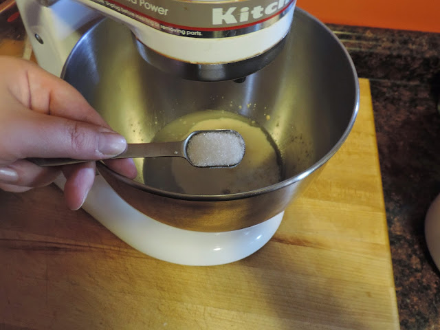 Sugar is being added to the yeast in the bowl of the Kitchenaid mixer.