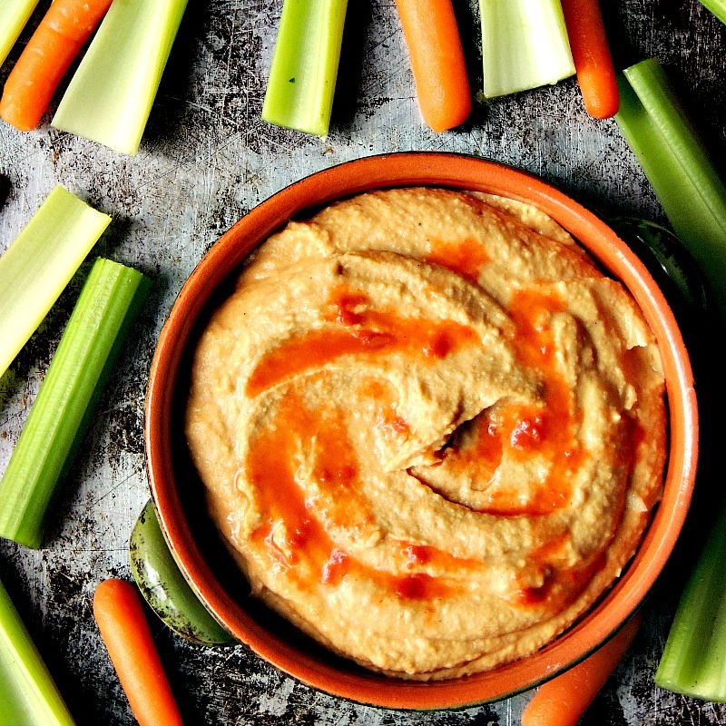 Buffalo Hummus in an orange and green bowl on a metal background with carrots and celery