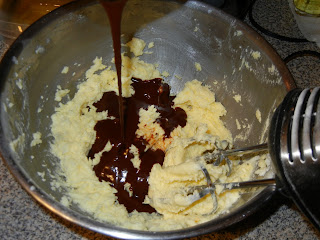 pouring chocolate into butter and sugar being beat together in a metal bowl with a hand mixer 