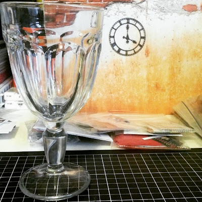 Empty wine glass sitting on a cutting mat in front of a selection of bagged kits and a one-twelfth scale miniature wall with a metal clock on it.