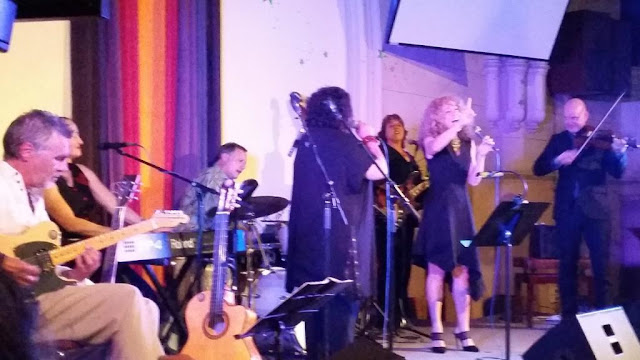 The complete ensemble with Vonda Last and Sandra Pires in black at centre stage. Around them in a semi-circle is a guitarist, keyboard player, drummer, electric bass player and violinist.  Sandra is raising her right hand as she is singing whle holding the microphone in her left. Vonda has a black and red bracelet around her left wrist as she raises the microphone to sing.