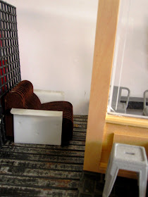 View through a mock up of the front of a modern dolls' house miniature cafe showing a lounge chair and several cafe chairs.