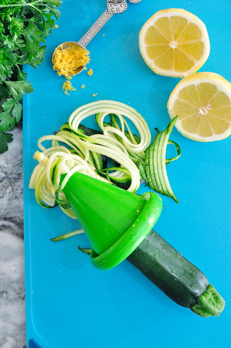 A shrimp scampi recipe with, zoodles, tomatoes and lemon create a healthy, low calorie weeknight meal. Parsley, spiralized zucchini, lemon zest and lemon on cutting board.