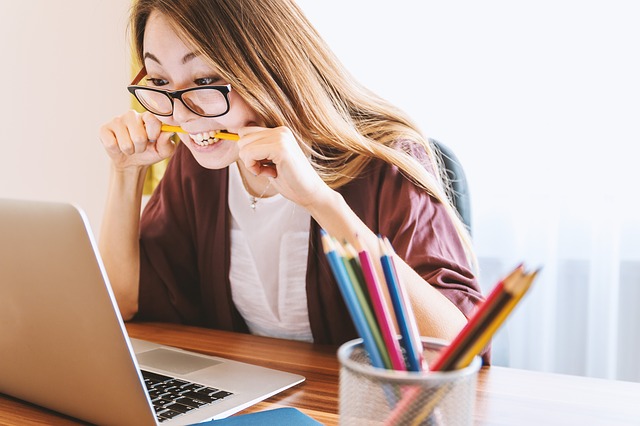 woman biting pencil while looking at laptop on desk