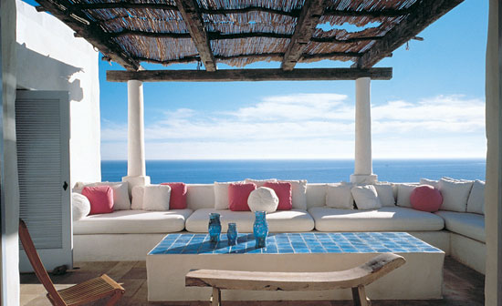 outdoor patio with white sectional sofa, red and white pillows, a block coffee table with blue tiles, reclaimed wood floors and a great view of the ocean