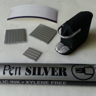 One-twelfth scale modern miniature 3D-printed Nespresso machine with silver highlights next to a silver caligraphy pen, three pieces of silver-coloured ribbed plastic and a photo of water. 