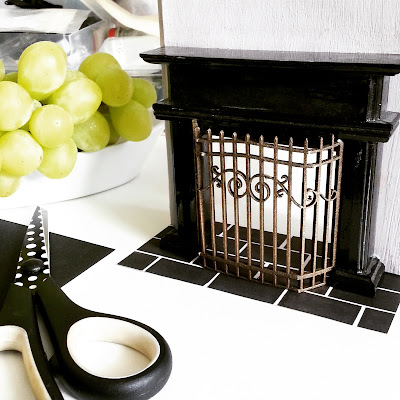 One-twelfth scale fireplace and chimney breast on a work table, with a brass.firescreen in front of it and a bowl of (life sized) grapes next to it.