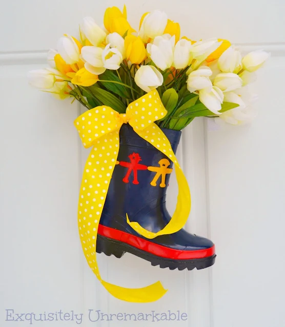 Small rubber rain boot on front door with tulips and yellow bow