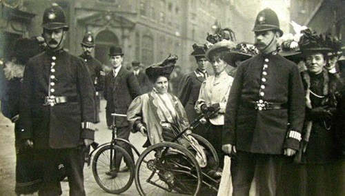 black and white photograph of Rosa May Billinghurst sitting in her adapted wheelchair after being arrested, surrounded by police officers and members of Women’s Social and Political Union