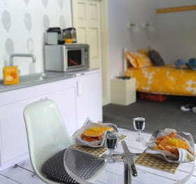 One-twelfth scale modern miniature motel room with a kitchenette at the front with a round table set for fish and chips and red wine. At the back of the room is a bed and bedside table.