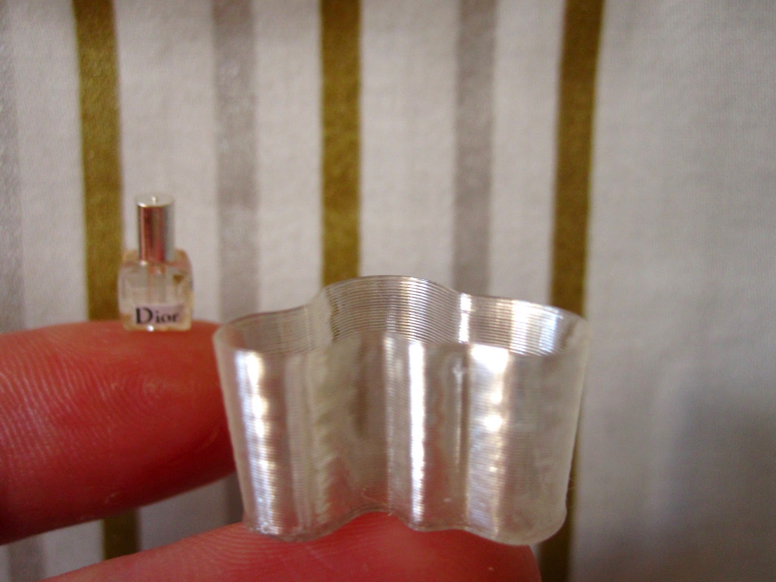 A miniature Dior perfume bottle and a miniature Aalto vase balanced on fingertips.