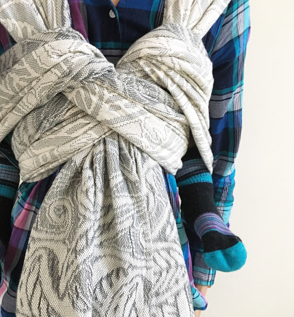 [Image of a woman’s torso featuring a gray botanical-patterned woven wrap carrier worn over a jewel-toned plaid top. Toddler on back has Mama’s wool socks on up to her knees to keep her feet and legs warm.]