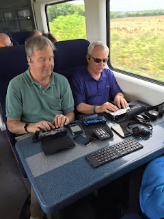 Mike and Bob on the train with their techonogy, BrailleNotes, phones, VictorReaders Stream