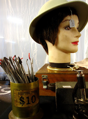 Corner of a market stall with a container of sculpting tools for sale, along with a mannequin head wearing a pith helmet, and a microscope.